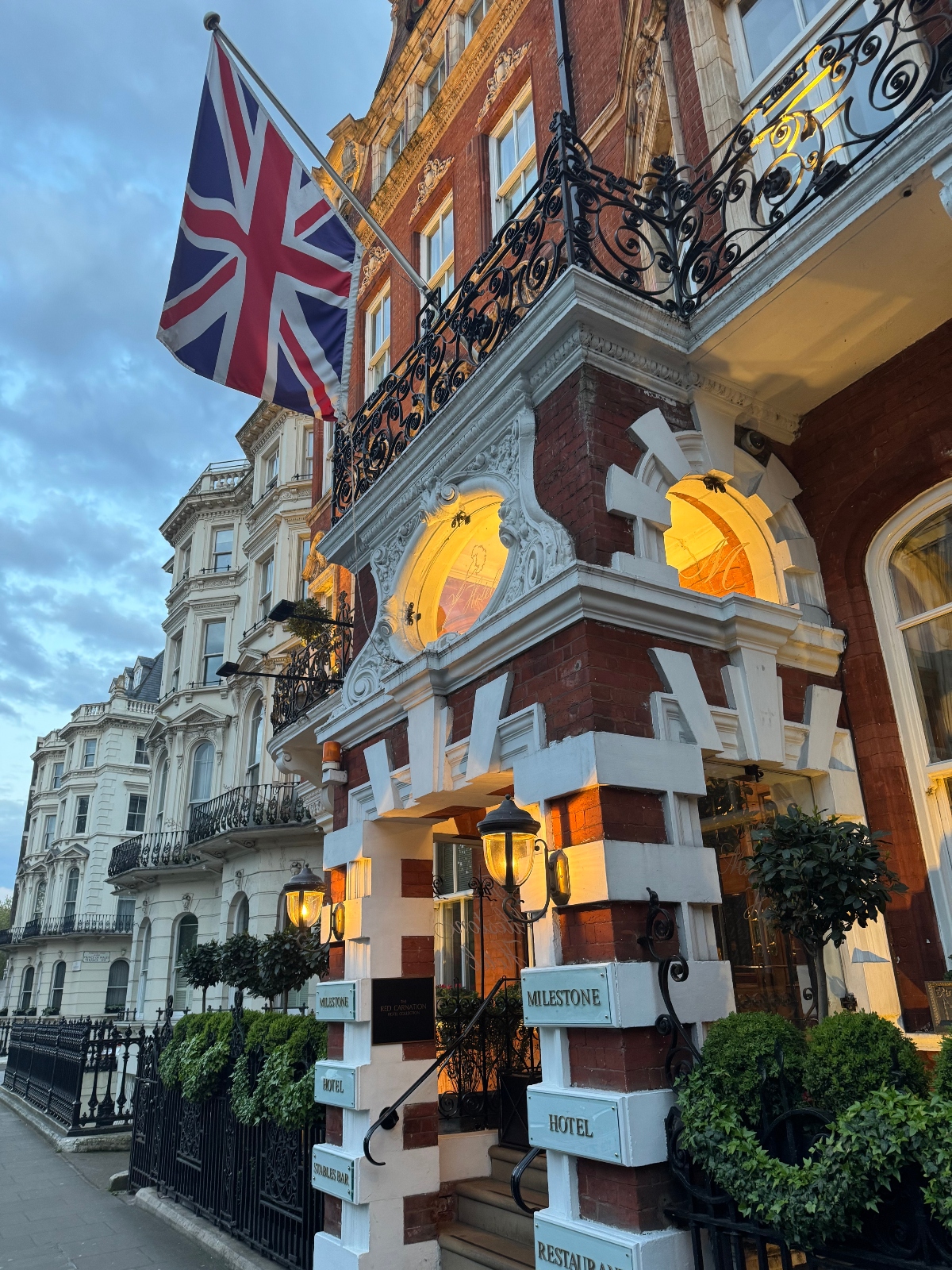 The exterior front entrance of the Milestone Hotel, among the top boutique hotels in London, glows with golden lights at dusk. The multi-story building is in a row of similar buildings; it is made of red brick with white moldings and a black wrought-iron balcony on the second floor. A Union Jack flag is mounted above the door and topiaries flank either side of the entrance.