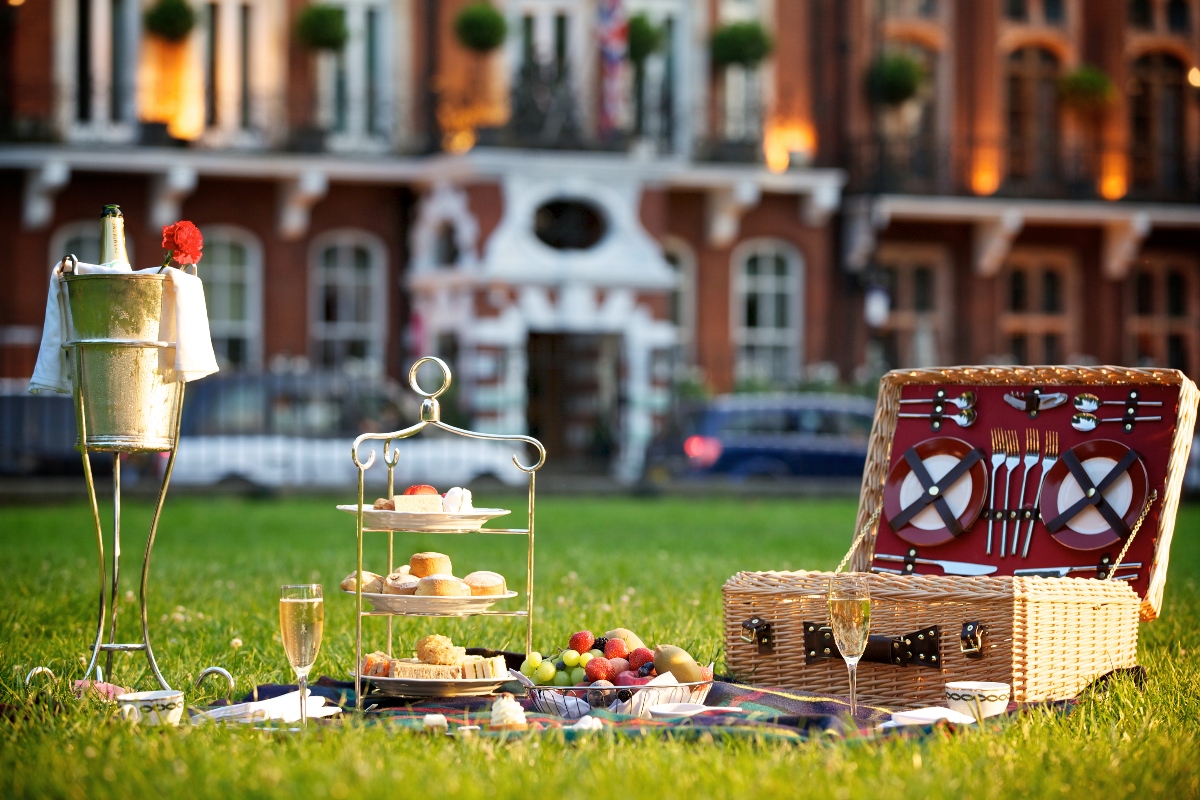 A luxury picnic setup on green grass features a wicker hamper with red interior compartments containing plates and utensils, alongside a champagne bottle in an ice bucket stand, a tiered serving tray with pastries and treats, champagne glasses, and fresh strawberries arranged on a tartan blanket. Historic brick buildings with Georgian architecture form the blurred background of this outdoor dining scene from The Milestone Hotel, one of the best boutique hotels in London.