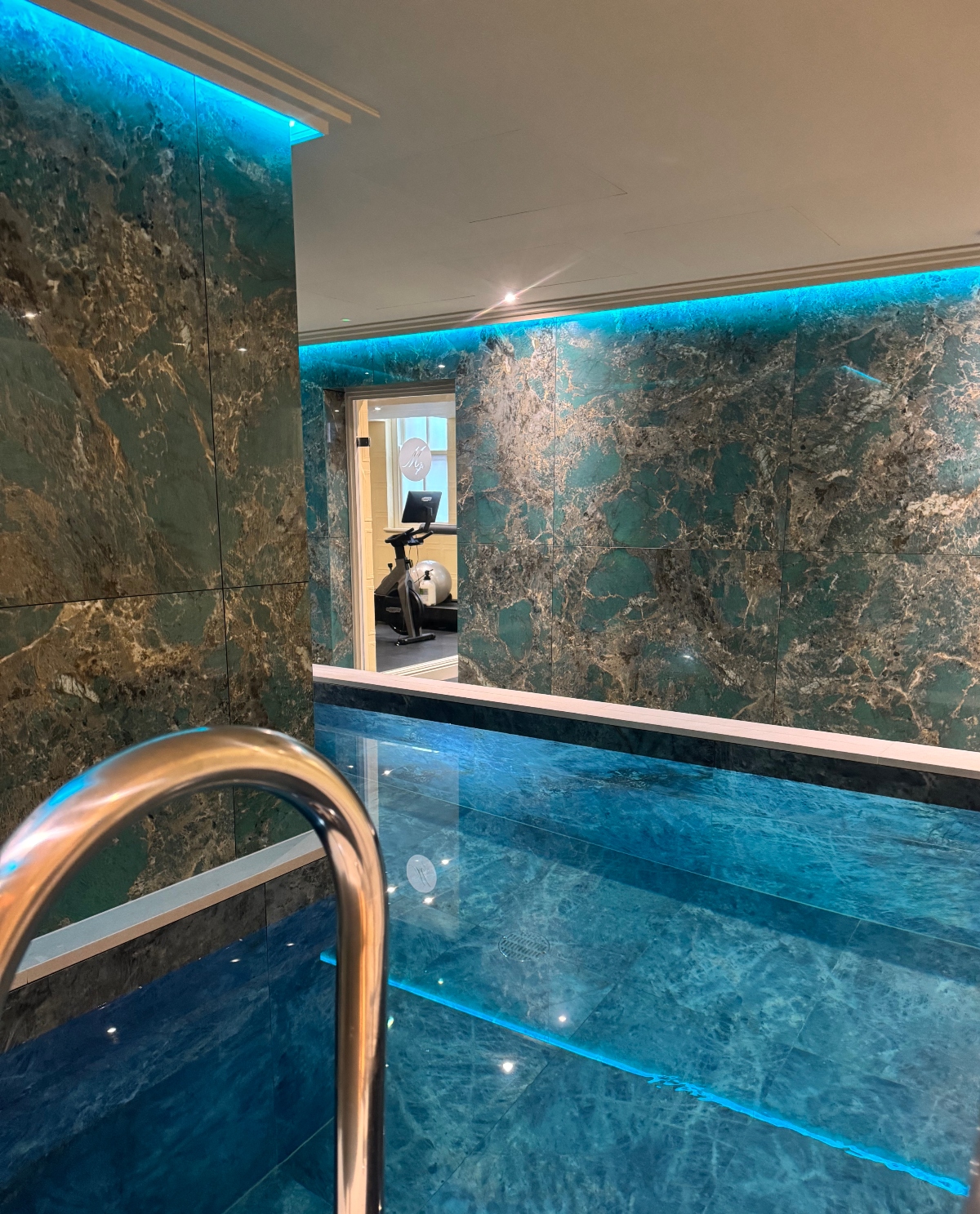 An indoor swimming pool features dark blue water surrounded by dark marble walls with gold veining, illuminated by blue LED strip lighting along the perimeter. A chrome handrail extends into the pool area, and through a doorway in the background, exercise equipment is visible in an adjacent fitness room with white walls.