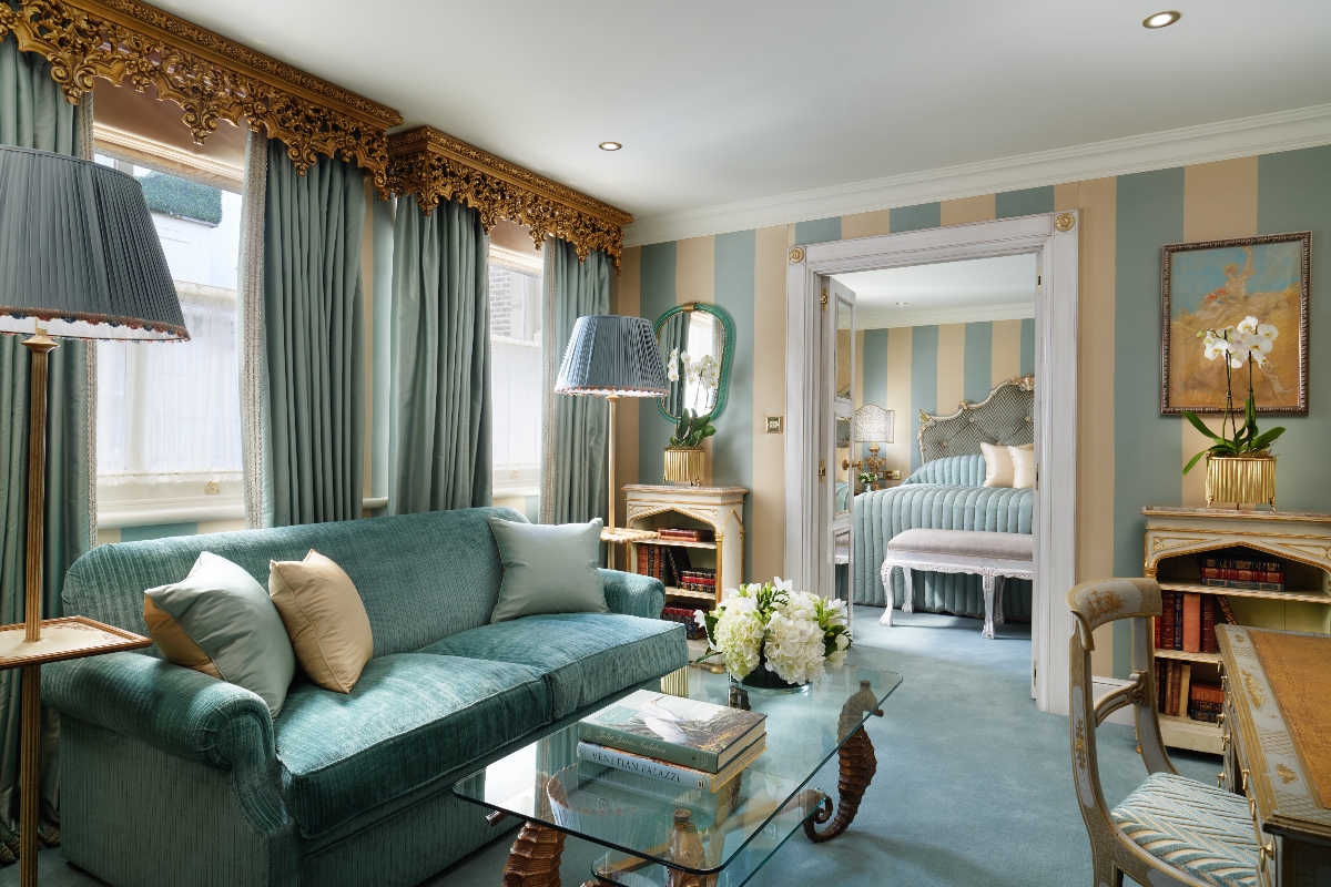 The opulent Venetian Suite at the Milestone Hotel features an elegant sitting room with a plush teal velvet sofa, ornate gilded curtain cornices, and sophisticated green and beige striped wallpaper that creates a refined English luxury atmosphere. The suite flows seamlessly from the living area into a glimpse of the bedroom beyond, showcasing classical furnishings, rich fabrics, and traditional European hotel grandeur with artwork and fresh floral arrangements.