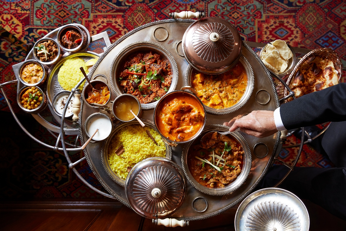 A large ornate silver serving tray displays numerous bowls containing various Indian dishes including yellow rice, orange and brown curries, flatbreads, and small accompaniments, with decorative covered serving dishes featuring ribbed lids. A person's hand in a dark suit sleeve reaches toward the elaborate spread, which is arranged on a richly patterned traditional carpet featuring red, blue, and gold geometric designs.