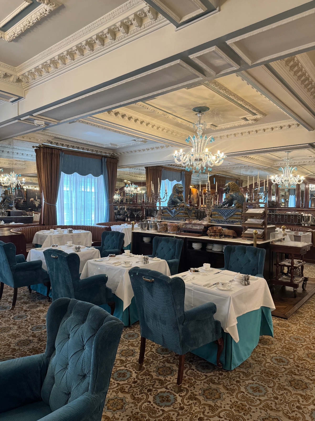 A formal dining room features multiple tables covered in white tablecloths surrounded by teal upholstered chairs, with an ornate coffered ceiling decorated with detailed moldings and crystal chandeliers. The space includes tall windows with brown curtains and blue inner panels, mirrored walls, decorative lion statues, and a patterned carpet covering the floor.