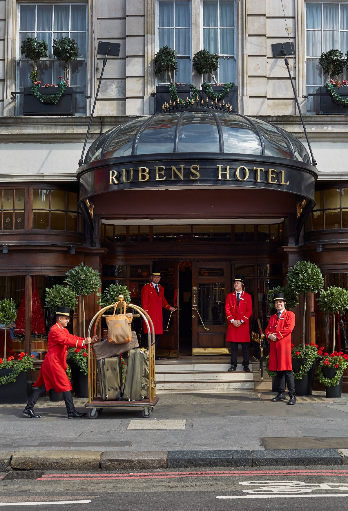 The elegant entrance of the Rubens at the Palace, one of the premier boutique hotels in London, features a distinctive curved glass canopy with the hotel name in gold lettering, flanked by several uniformed doormen in traditional red coats and caps providing attentive service. One doorman pushes a brass luggage cart while his colleagues stand ready to assist guests, creating a classic scene of British hospitality against the backdrop of the hotel's refined Georgian-style facade decorated with topiary plants and seasonal flowers.