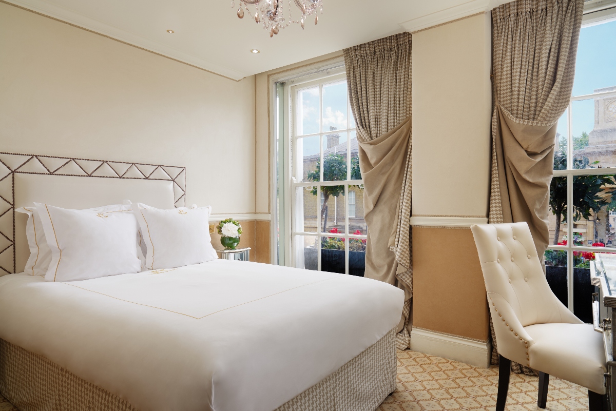 An elegant Royal Mews View room at the Rubens at the Palace features a luxurious bed with crisp white linens and a distinctive geometric-patterned headboard, complemented by a plush cream upholstered chair and crystal chandelier overhead. The room showcases sophisticated traditional decor with elaborate patterned curtains adorned with decorative swags framing tall windows that offer views of the historic Royal Mews area.