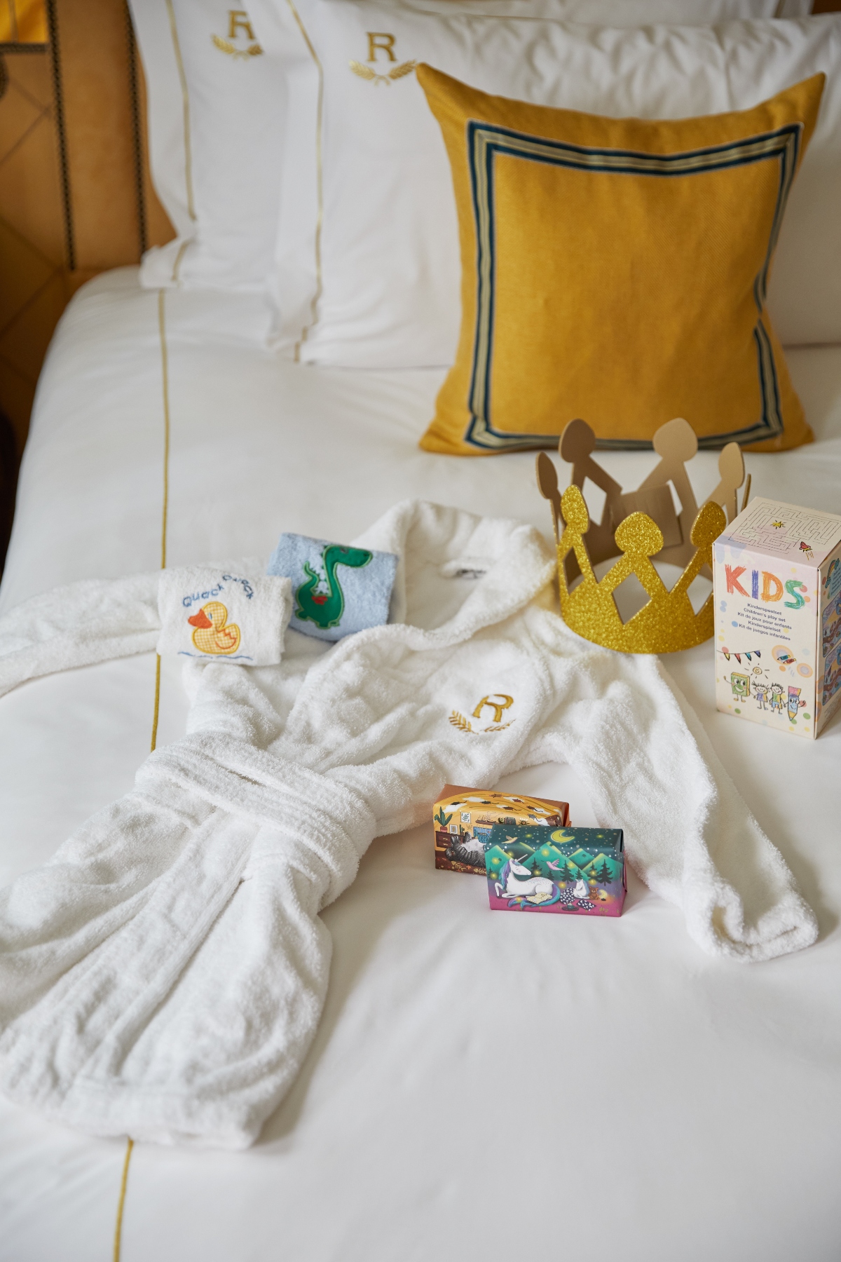 A white hotel bed displays children's amenities including a small white terry cloth bathrobe with gold embroidered logo, a golden paper crown, colorful gift boxes, and small towels with duck and animal designs. The items are arranged on white linens alongside a yellow decorative pillow with dark trim and a "KIDS" branded card or pamphlet.