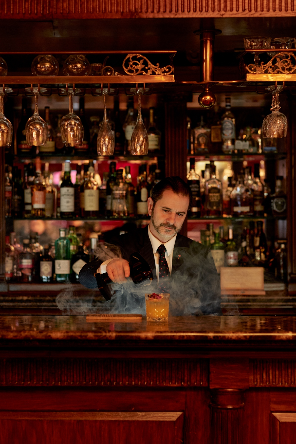 A skilled bartender in a dark suit and tie creates a dramatic cocktail with billowing smoke at the elegant New York Bar at Rubens at the Palace, working behind a richly appointed mahogany bar. The sophisticated setting features an extensive collection of spirits displayed on illuminated shelves, hanging copper-toned wine glasses, ornate woodwork, and warm ambient lighting.