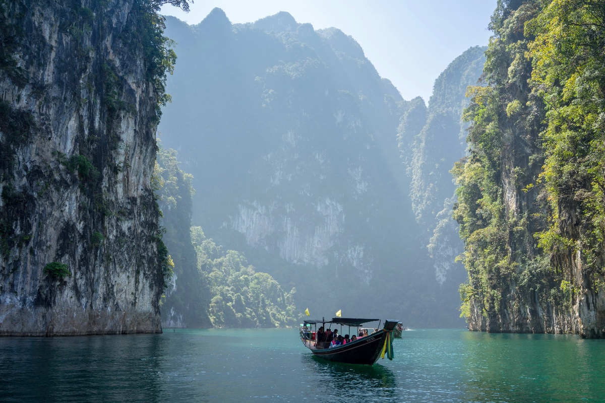 A traditional Thai boat glides through turquoise waters in the shadows of tall karst cliffs covered with vegetation. 