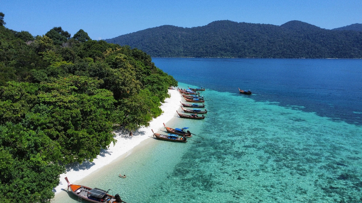 Traditional Thai boats sit on a crescent of white sand bordered with lush tropical foliage and shallow turquoise water on the island of Koh Lipe, one of the best remote Thailand vacation destinations. The blue ocean and green hills sit in the distance. 