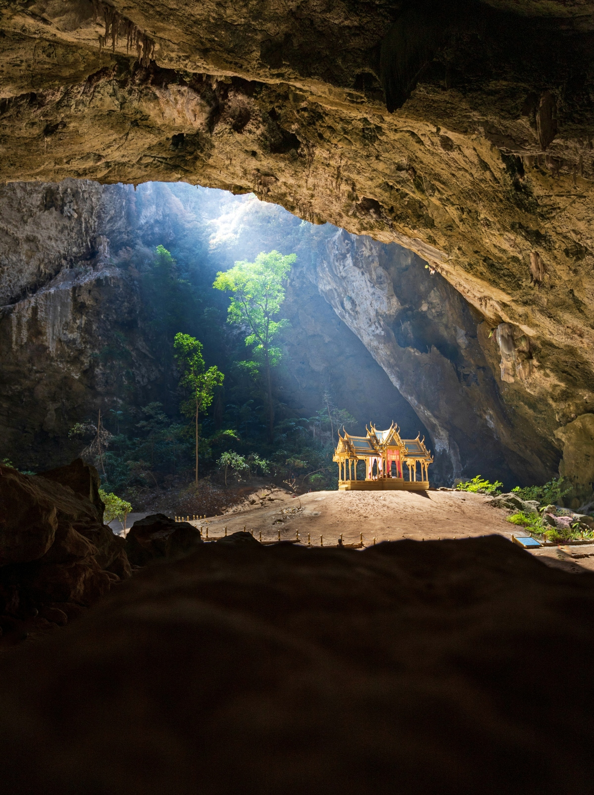 A golden temple stands at the opening to Phraya Nakon Cave, one of the best off-path Thailand vacation destinations, seen from inside the cave itself. It is dwarfed by the curving rock formations in the foreground. A shaft of sunlight illuminates the temple and two leafy green trees behind it. 