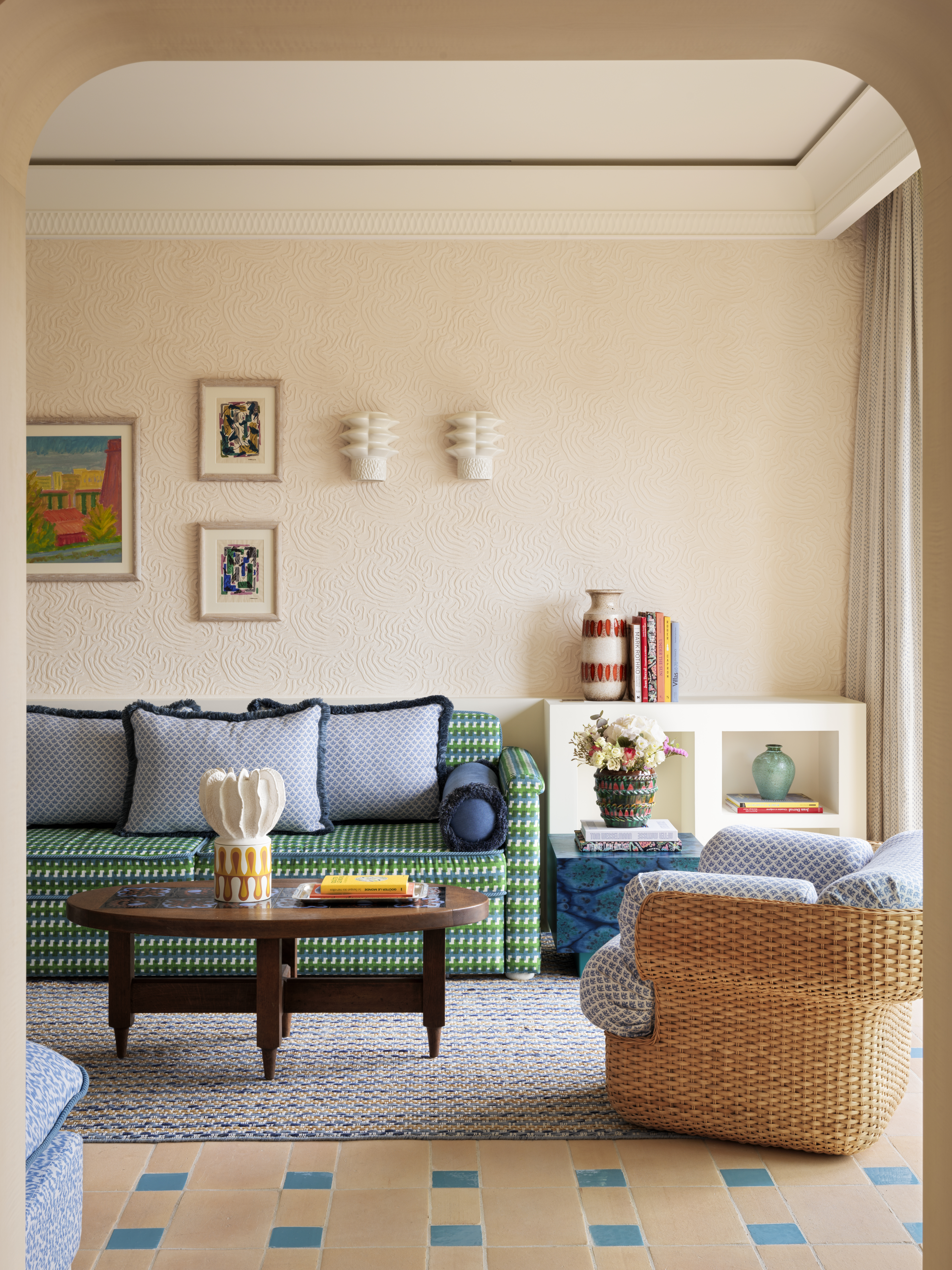 This interior features a curved alcove with built-in seating upholstered in green and white geometric fabric, accompanied by blue and patterned throw pillows, a dark wooden round coffee table, and a wicker armchair. The space includes built-in shelving displaying books and decorative objects, framed artwork on textured wallpaper, and a geometric tile floor in blue and beige tones.
