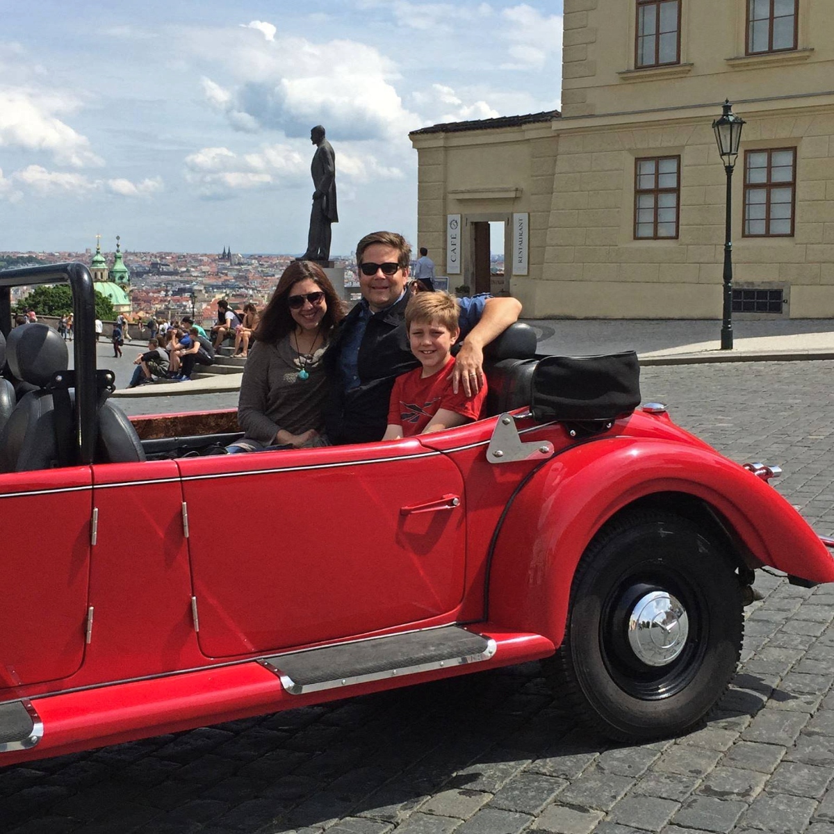 Three people, including two adults and a child, sit in a red vintage convertible car parked on a cobblestone plaza in what appears to be a historic European city. Behind them stands a classical stone building and a statue on a pedestal, with a cityscape and cloudy sky visible in the background.