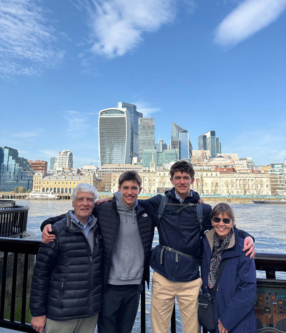 Four people pose together for a photo on a waterfront walkway by the River Thames in London, with their arms around each other in a group formation. Behind them is a city skyline featuring modern glass skyscrapers and historic buildings across a body of water, under a partly cloudy blue sky.