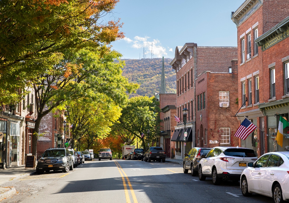A tree-lined street runs through a historic downtown area with red brick buildings on both sides, cars parked along the curbs, and American flags displayed on several storefronts. In the background, a church steeple rises above the buildings with forested hills visible under a clear blue sky, while autumn foliage creates a canopy of green and golden leaves over the street.