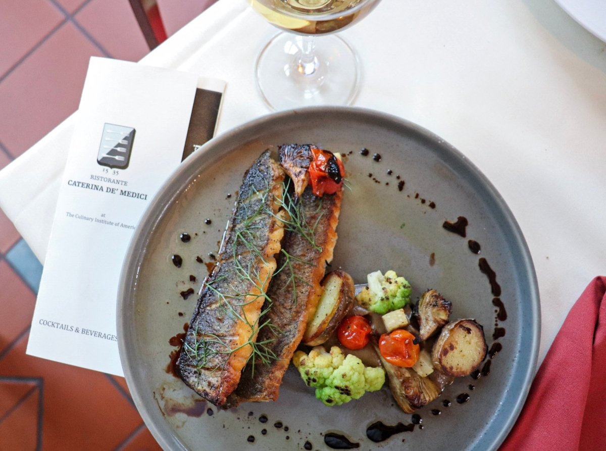 A gray ceramic plate contains a seared piece of fish garnished with fresh herbs, accompanied by roasted vegetables including cauliflower, mushrooms, and cherry tomatoes, with dark sauce dots arranged around the plate. The dish is photographed on a white table alongside a wine glass and a menu or document labeled "Caterina de' Medici."