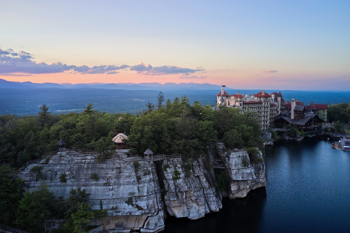 A large resort complex with multiple buildings of varying architectural styles sits atop dramatic limestone cliffs overlooking a lake, surrounded by dense forest. The scene is captured during golden hour with layered mountains visible in the background and a partly cloudy sky creating warm lighting across the landscape.