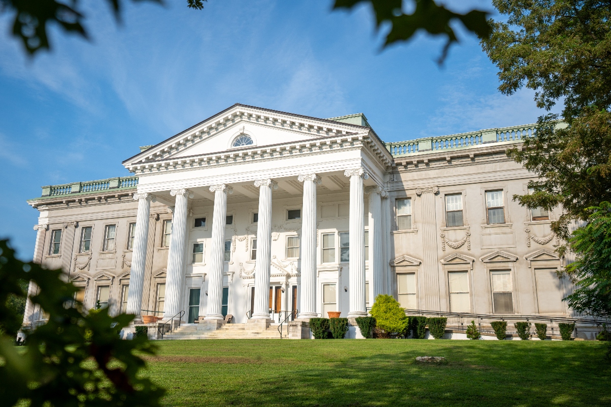 A large neoclassical building with white stone construction features a prominent portico supported by six tall Corinthian columns and topped with a triangular pediment. The symmetrical structure sits on manicured green lawns with landscaped shrubs, framed by tree branches under a clear blue sky.