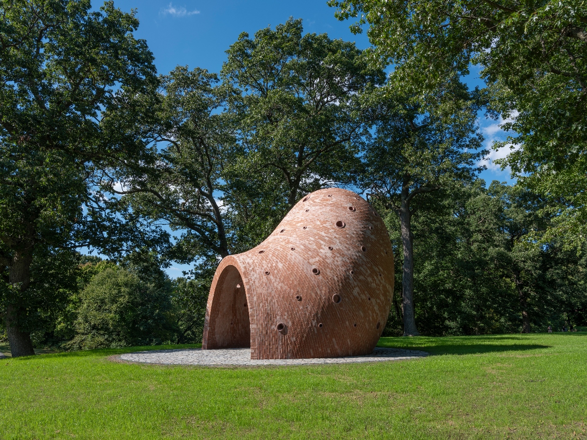 A large, curved brick structure with an organic, pod-like shape sits on a stone platform in a grassy park setting, featuring circular openings of various sizes scattered across its surface and a large arched entrance at one end. The red-orange brick installation is surrounded by mature green trees under a blue sky with white clouds.