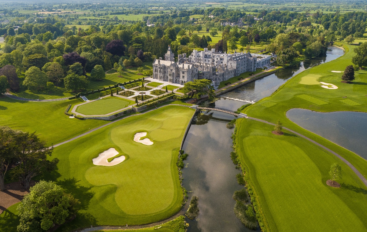 Aerial view shows a large rock castle or manor house with towers and formal architectural features surrounded by formal gardens with geometric hedge patterns. Located next to the Champions Golf Course, the property has multiple holes with sand pits, water hazards and lush green fairways that wind through the landscape. A river bends at the scene, while vast countrysides with fields, trees and rolling terrain extends to the cloudy horizon.