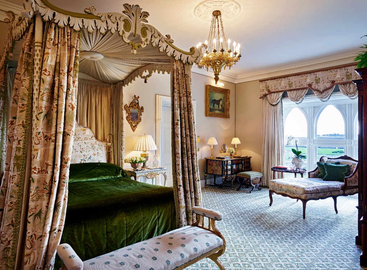 A luxurious bedroom features a four-holding bed with beautifully carved canopy, floral chintz curtains and emerald green velvet bedding. The room is decorated in the traditional English country house style with floral wallpaper, crystal chandeliers, period furniture including lounge chairs and large arched windows with ornate curtains. Antique furniture, desk lamps and decorative artwork complete the luxurious interior on the patterned carpet.