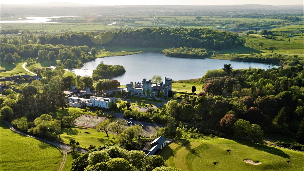 Aerial view shows a medieval castle with towers and walls on walls located near a wide lake surrounded by wooded hills. The castle complex includes modern hotel or resort buildings as well as historic structures, manicured lawns and golf courses featuring sand castles and fairways in the foreground. The scenic rural landscape extends to the distance, rolling fields, dense woodlands and pastoral pastoral pastoral stretches to the distance under soft atmospheric lighting.