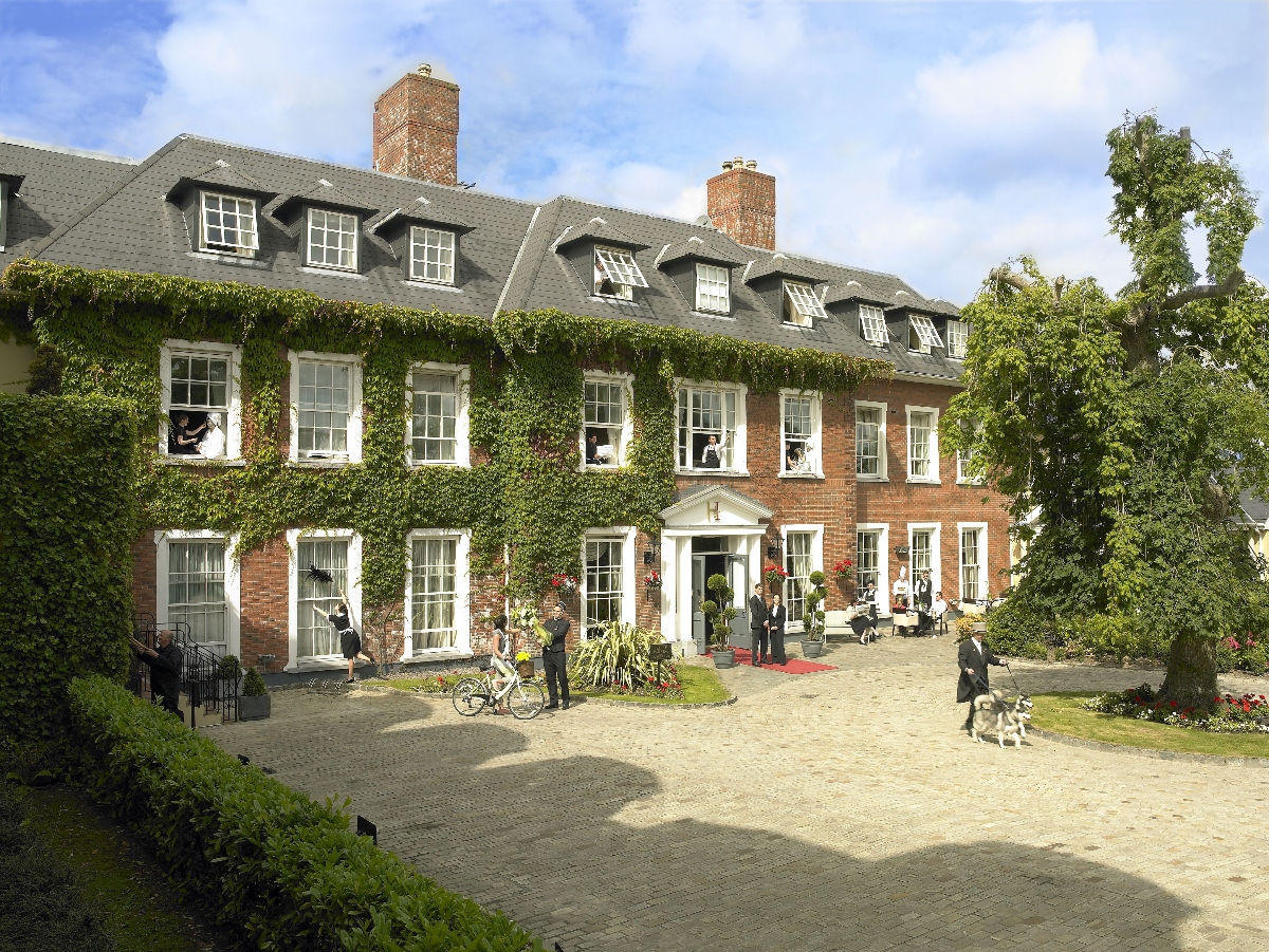 A large Georgian style brick mansion with ivy-covered walls, with multiple stories, windows and famous brick chimneys under partially cloudy skies. The entire windows of the building feature white framed windows with a central entrance to white columns, while the curved gravel courtyard in front offers several people and dogs to see. Mature trees and landscaping surrounding the property, bikes are located at the entrance.