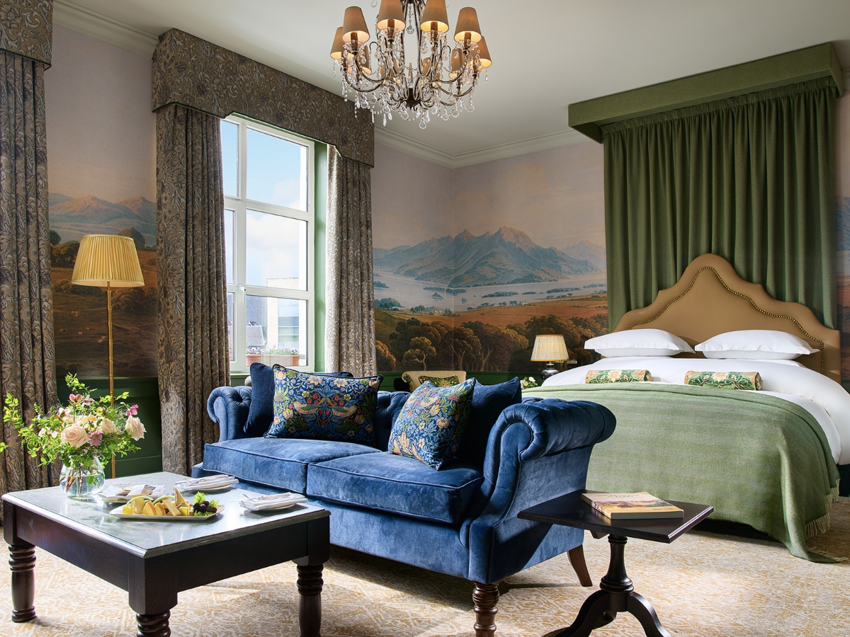 A luxurious hotel suite features a bed with white linen and beige upholstered headboard next to a blue velvet sofa with patterned throwing pillows on a dark wood coffee table. The room is decorated with large murals that depict the mountain and rural scenes on the walls, green curtains and patterned curtain frames. Crystal chandeliers with fabric lampshades hung from the ceiling, illuminating the traditionally appointed space with crowns and refined furniture.
