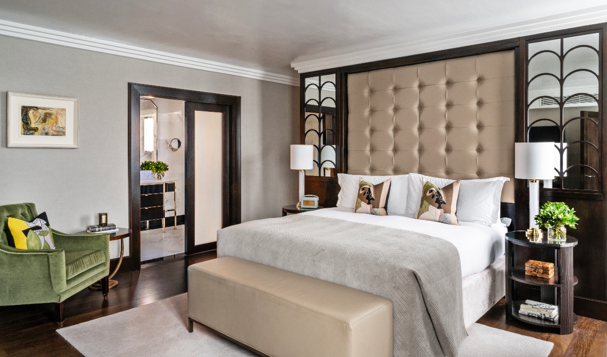 A luxurious bedroom features a king-size bed with white linen and a large tufted beige headboard framed by deep wood and decorative arched panels. The room includes green velvet armchairs, beige leather benches at the foot of the bed, and matching bedside tables, and white table lamps on the black hardwood floors. An open doorway to the left leads to the adjacent room, while neutral grey walls and crown molding complete the exquisite interior design.