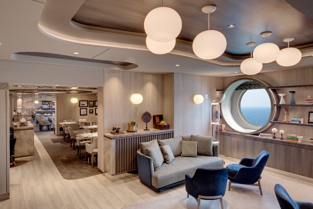 A modern interior lounge features a gray sectional sofa and blue accent chairs arranged around a central area, with multiple large spherical pendant lights hanging from a curved ceiling with wood paneling. The space includes built-in shelving with books and decorative objects, a large circular porthole window showing an ocean view, and an adjacent dining area with tables and chairs visible in the background.