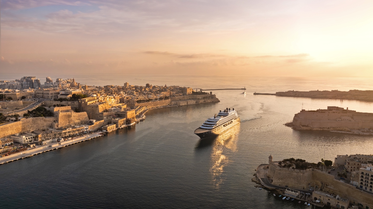 A large white cruise ship navigates through a harbor surrounded by historic golden stone fortifications and buildings during golden hour lighting. The aerial view shows a Mediterranean-style port city with distinctive limestone architecture, defensive walls, and breakwaters extending into calm waters under a soft, peachy sky.