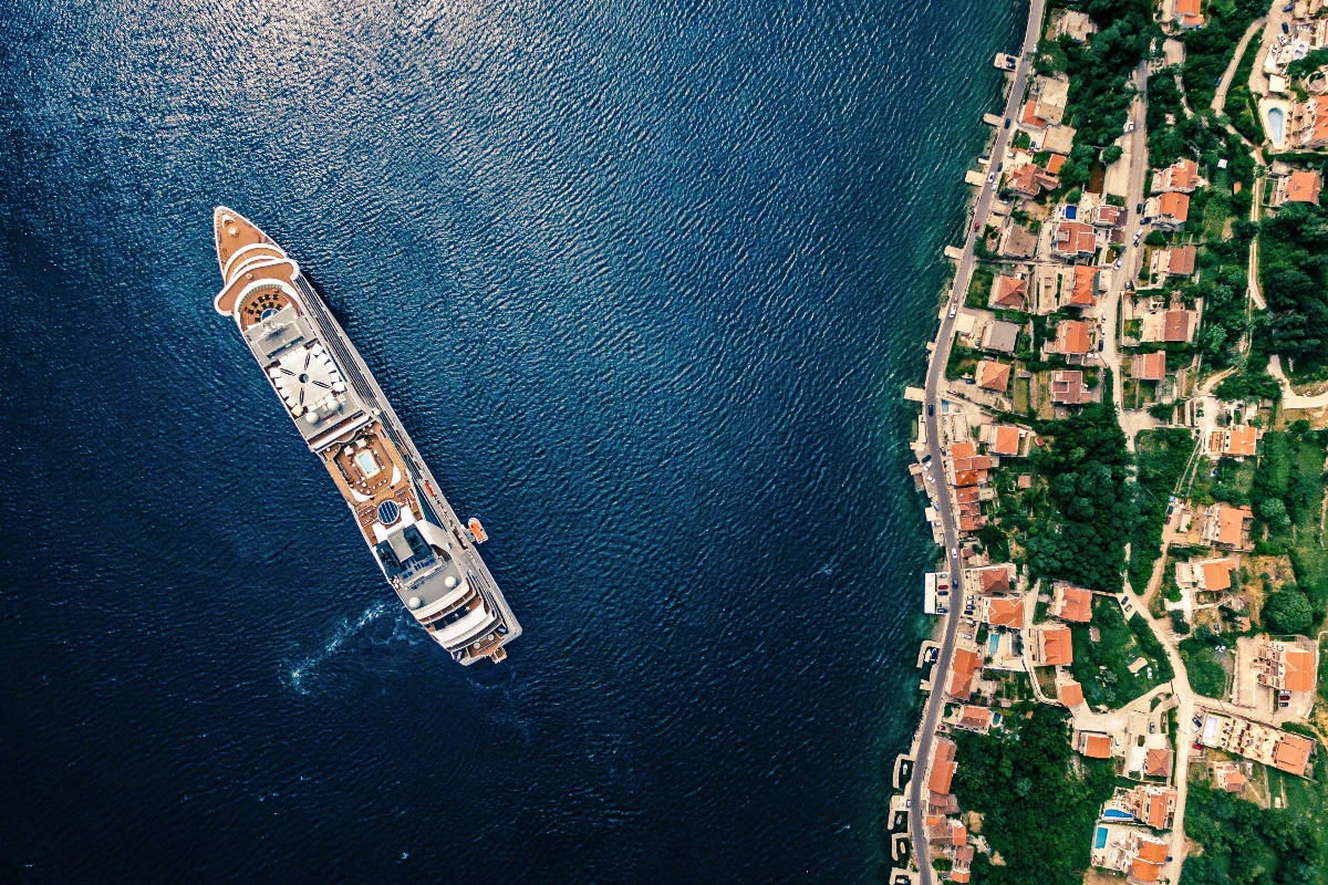 An aerial view shows a large white cruise ship or yacht anchored in deep blue water near a coastal settlement. The shoreline features a cluster of red-roofed buildings and houses nestled among green vegetation, connected by winding roads and pathways.