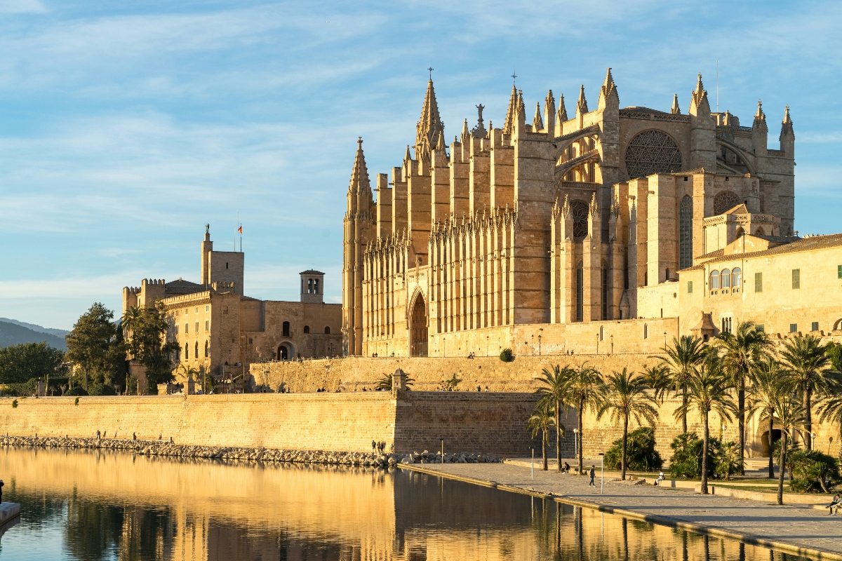A large Gothic cathedral with multiple spires and ornate stonework dominates the skyline, situated on elevated ground above a waterfront with defensive walls and palm trees. The golden-colored stone buildings are reflected in the calm water below, while the warm lighting creates a luminous effect on the cathedral's facade and surrounding architectural complex.