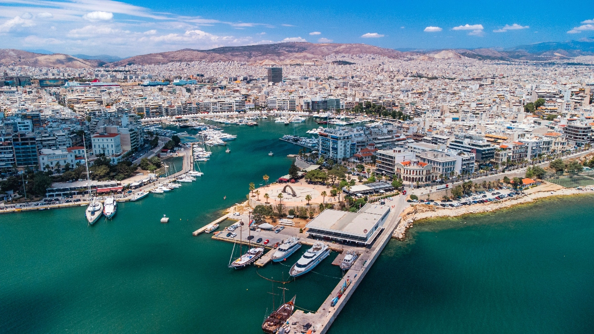 An aerial view shows a large marina filled with numerous white boats and yachts, centered around a prominent concrete pier that extends into turquoise-colored water. The harbor is surrounded by white and light-colored multi-story buildings that spread across the landscape toward barren hills in the background, with palm trees and waterfront promenades visible along the shoreline.