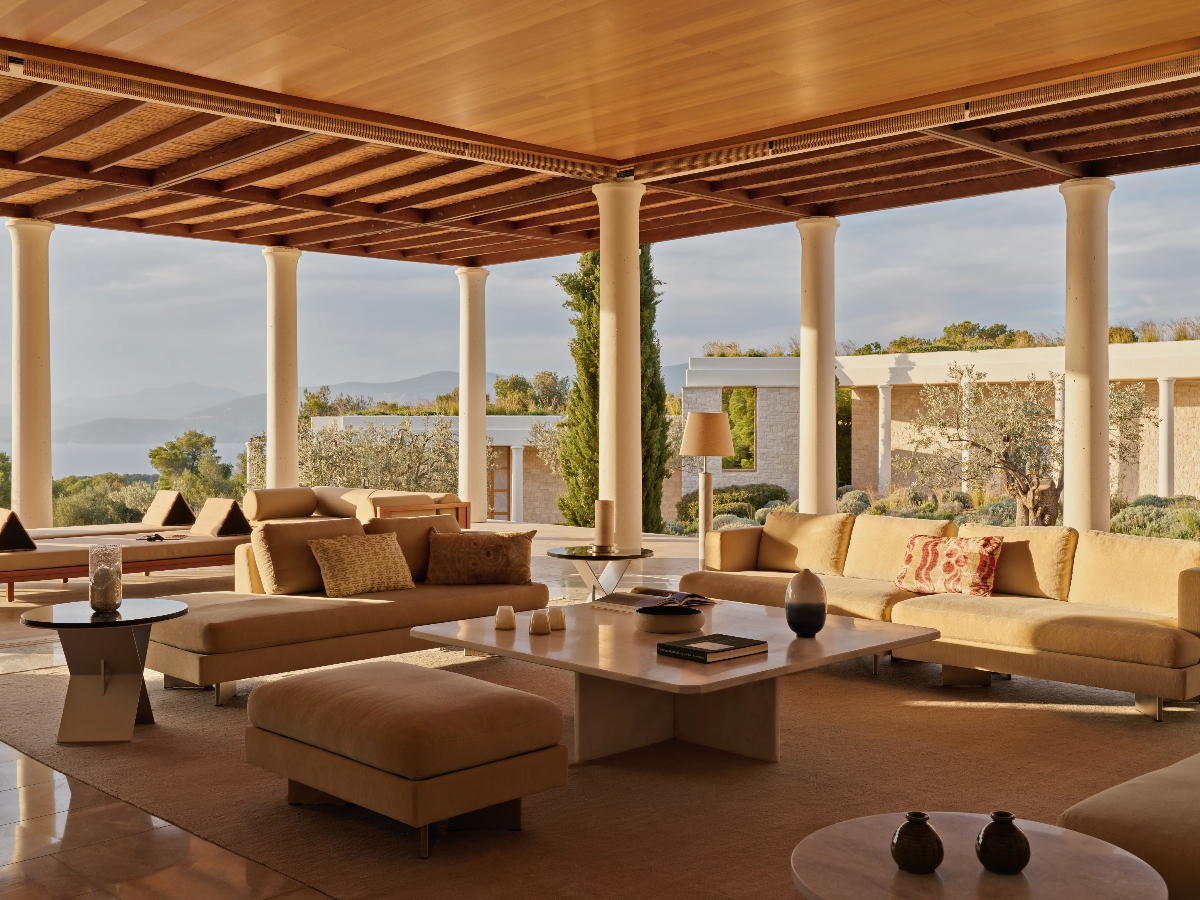 A covered outdoor terrace features multiple cream-colored sofas and seating areas arranged around low coffee tables, supported by white columns and a wooden slatted ceiling structure. The open-air space overlooks a landscape with modern buildings, trees, and distant mountains visible through the columned openings.