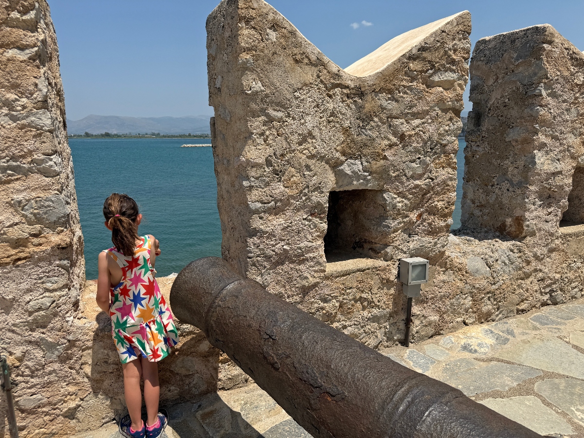 A young girl in a colorful star-patterned dress stands among ancient stone ruins with weathered limestone walls and archways, with a small informational sign visible nearby. The archaeological site overlooks a blue coastal vista with distant mountains visible across the water under a clear sky.