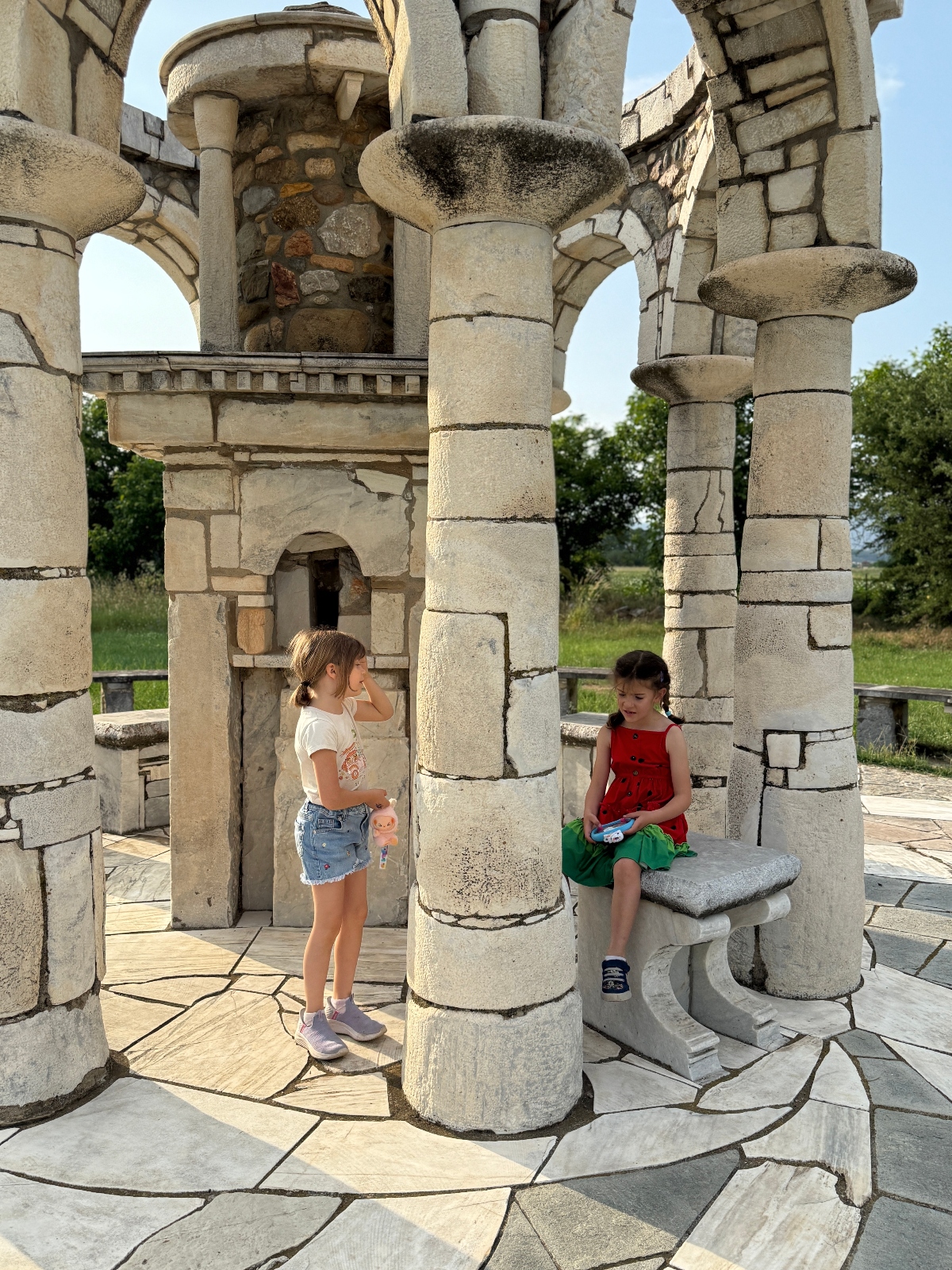 Two young children explore ancient stone ruins featuring tall columns with ornate capitals, arched openings, and detailed masonry work. One child in a white shirt and denim shorts stands near a fluted column while another child in a red dress sits on a carved stone bench, surrounded by the weathered limestone architecture and patterned stone flooring.