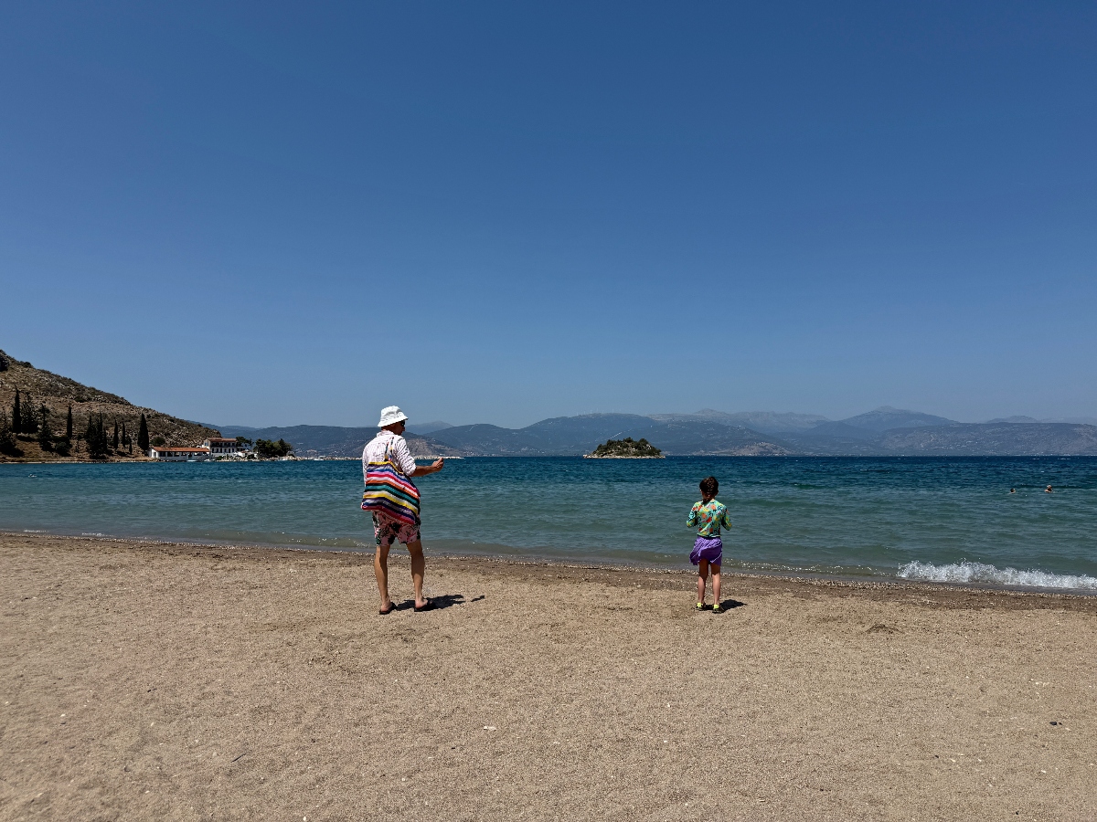 A person wearing a white hat and colorful striped clothing stands on a sandy beach alongside a smaller figure, both facing the calm blue water. The beach is set in a scenic bay surrounded by mountainous terrain under a clear blue sky, with distant buildings visible along the coastline and a small island in the water.