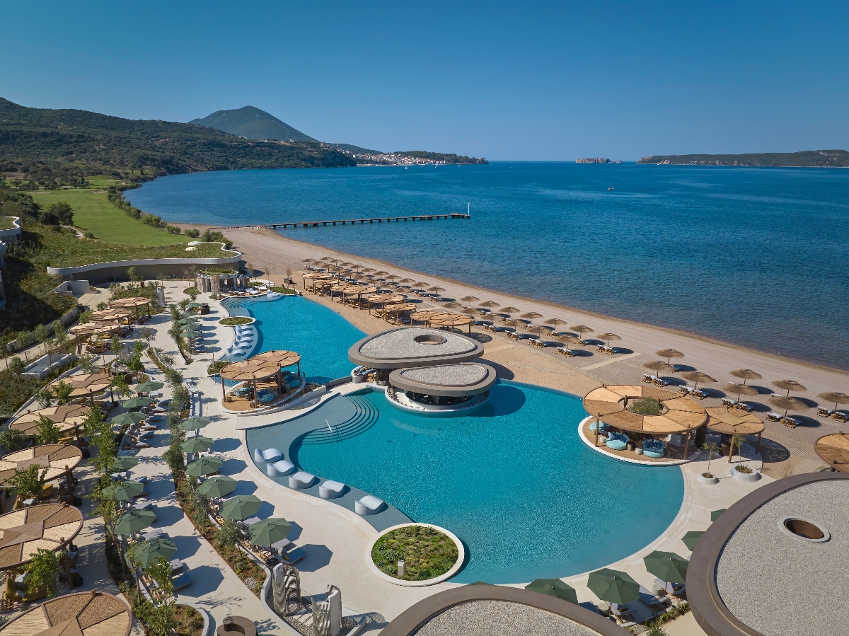 This aerial view shows a large, curved swimming pool complex situated directly on a sandy beach, with the pool featuring circular island structures and surrounded by white lounge chairs and umbrellas. Beyond the beach area, calm blue waters extend toward mountainous coastline in the distance, with a wooden pier extending into the water.