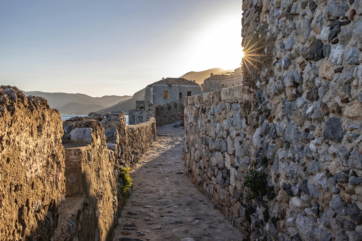 A narrow stone-paved walkway runs between weathered stone walls, with bright sunlight creating a starburst effect from behind the ancient masonry. In the background, white buildings are visible against distant mountains, while the golden lighting illuminates the rough-textured stone blocks and cobbled pathway.