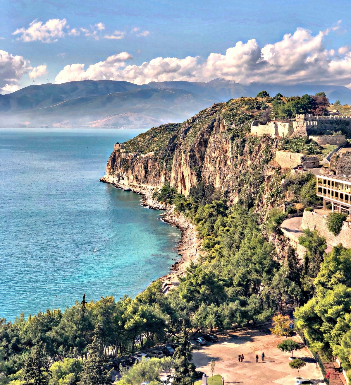 A dramatic coastal landscape shows steep limestone cliffs covered in green vegetation rising from bright turquoise water, with ancient stone fortifications and castle ruins perched on the clifftops. The scene includes a paved viewing area at the bottom where small figures of visitors can be seen, while distant mountains are visible across the water under a partly cloudy sky.