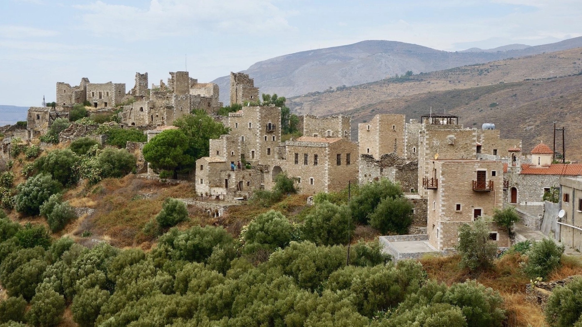 A hillside settlement features numerous stone buildings of varying states of preservation, from complete ruins to partially intact multi-story structures, built with light-colored limestone blocks. The terraced landscape is dotted with green vegetation and shrubs, set against a backdrop of arid mountains under a partly cloudy sky.