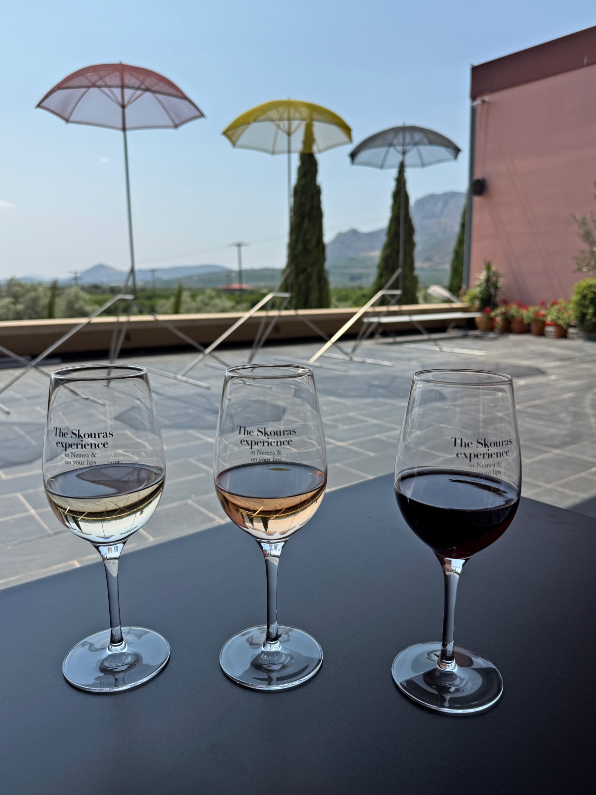Three wine glasses labeled "The Skouras experience" sit on a table, containing white, rosé, and red wine respectively. The scene is set on an outdoor terrace with colorful umbrellas, lounge chairs, and a mountainous landscape visible in the background under a clear blue sky.
