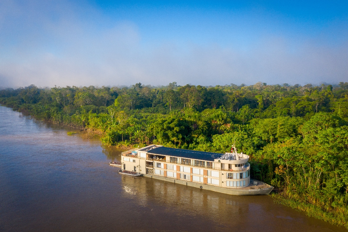 A white multi-deck river cruise vessel is anchored on a wide, muddy-brown river surrounded by dense tropical rainforest. The boat features multiple levels with windows and appears designed for passenger accommodations, positioned near the heavily forested shoreline. The scene is set under a hazy sky with lush green canopy extending as far as the eye can see along both sides of the waterway.