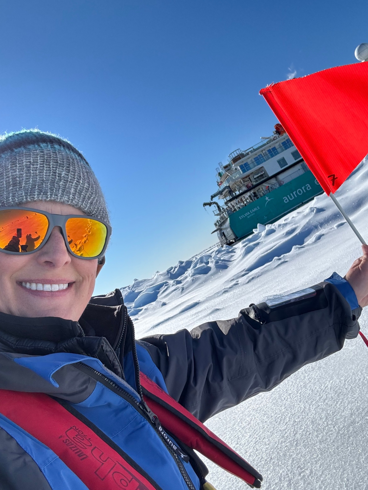 A person wearing orange-tinted sunglasses, a knit hat, and colorful winter expedition gear takes a selfie while holding a small red flag in a snowy polar landscape. Behind them, an expedition cruise ship with "aurora" visible on its hull is positioned among snow-covered terrain under a clear blue sky. The scene appears to be set in an Arctic or Antarctic environment with pristine white snow extending across the frame.