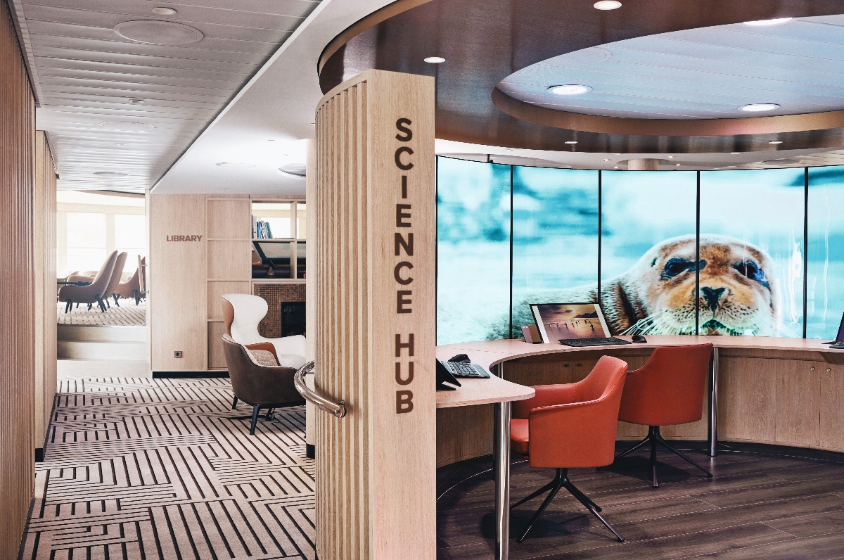 A modern interior space labeled "SCIENCE HUB" features workstations with orange chairs positioned at desks with computer equipment, set against large windows displaying an underwater view of what appears to be a seal or marine mammal. The contemporary design includes wood paneling, geometric patterned flooring, and curved ceiling elements with recessed lighting. In the background, a library area with seating can be seen, creating a multi-functional research and study environment.
