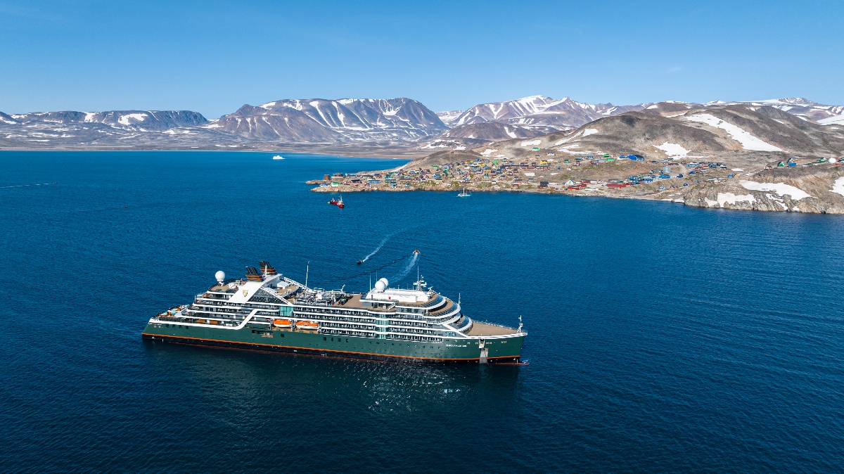 An aerial view shows a large cruise ship anchored in deep blue waters near a remote coastal settlement. The settlement consists of colorful buildings scattered across barren, rocky terrain with patches of snow, situated on a peninsula or coastal area. Snow-capped mountains rise dramatically in the background under a clear blue sky, creating a stark Arctic or sub-Arctic landscape.