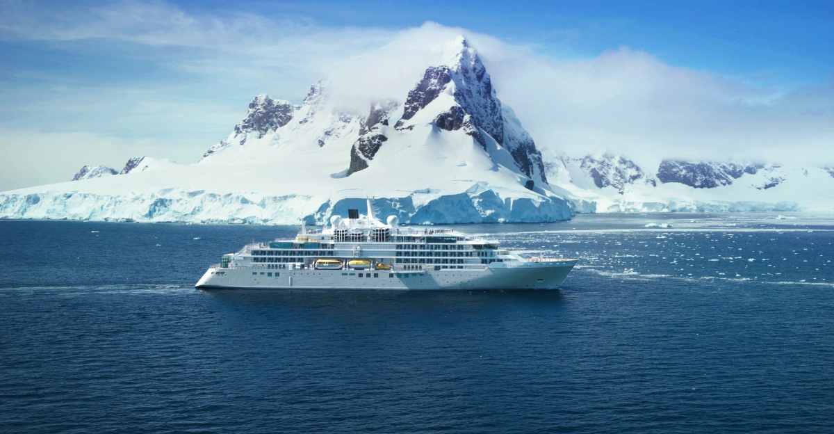 A white cruise ship navigates through dark blue Antarctic waters with a dramatic backdrop of snow-covered mountains and glacial formations. The towering peaks rise sharply from the water, partially shrouded in clouds, while icebergs and glacial ice can be seen along the coastline. The scene captures the pristine wilderness of what appears to be the Antarctic Peninsula under a partly cloudy sky.