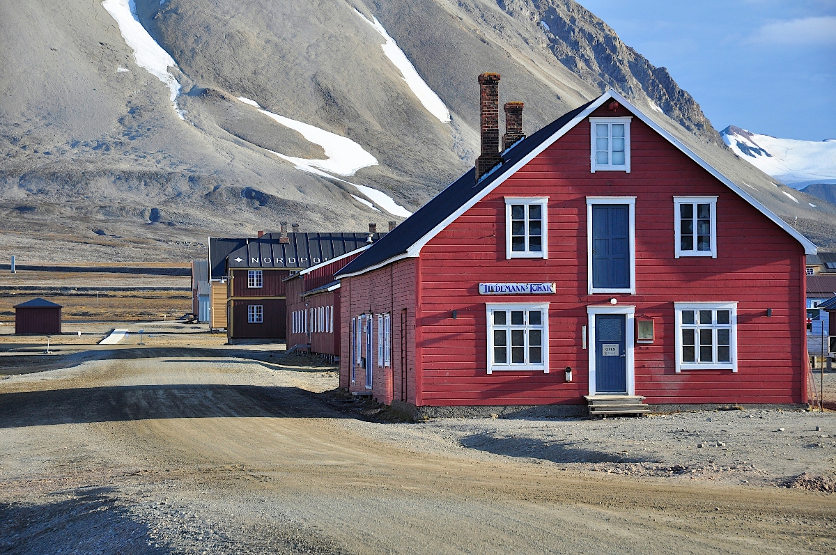 A bright red wooden building with white trim and blue doors stands prominently in the foreground of what appears to be an Arctic settlement. Behind it are several other colorful wooden structures, including a dark building marked "NORDPOL," all situated on unpaved ground. The buildings are set against a dramatic backdrop of snow-capped mountains and barren, rocky terrain under a clear blue sky.