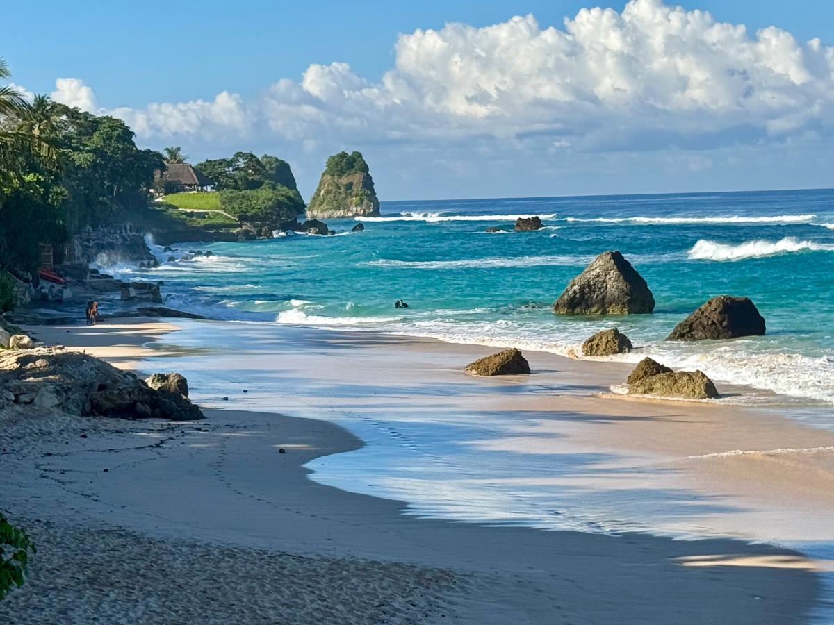 A tropical beach scene shows a curved sandy shoreline with turquoise waves washing ashore and several large rocks scattered across the beach and in the shallow water. Lush green cliffs covered in tropical vegetation, including palm trees, rise dramatically from the coastline under a blue sky filled with white clouds.