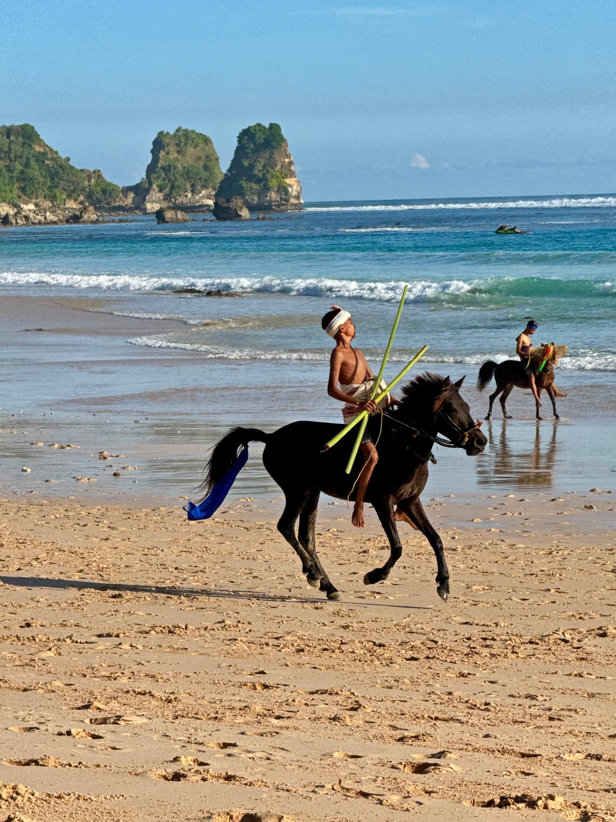 A person rides a dark horse on a sandy beach while holding long yellow-green poles, with ocean waves rolling onto the shore in the background. The scenic coastal setting features distinctive vegetation-covered rock formations rising from the turquoise water and additional horseback riders visible in the distance.