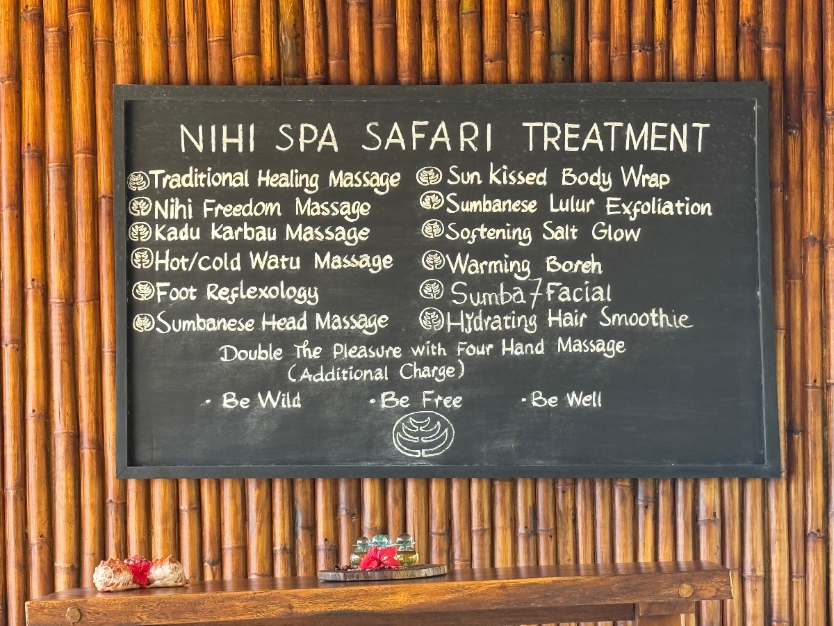 A black chalkboard sign mounted on a bamboo wall displays "NIHI SPA SAFARI TREATMENT" as the heading, followed by a menu of various massage and spa services including traditional healing massage, reflexology, body wraps, and facial treatments. Below the sign is a wooden shelf holding decorative items including a small woven basket with red flowers and a glass container.